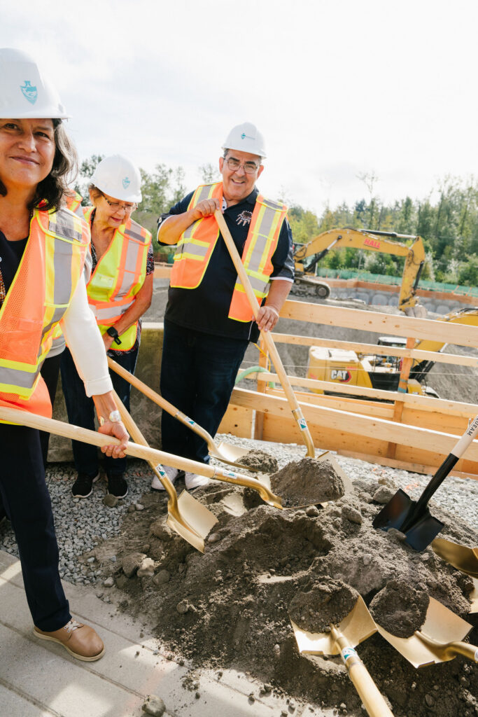 Attendees in hard hats and safety vests turning soil with ceremonial golden shovels at the East Wind groundbreaking, with excavation equipment behind them.