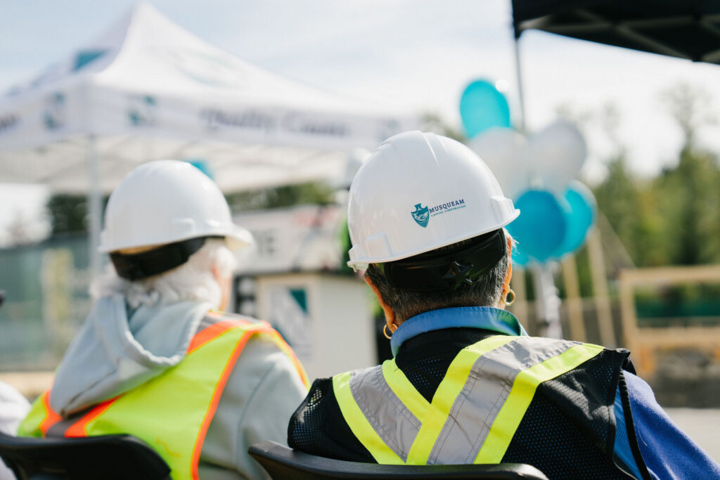 Two attendees seen from behind in hard hats and safety vests at the groundbreaking ceremony, one hard hat bearing the Musqueam Capital Corporation logo.