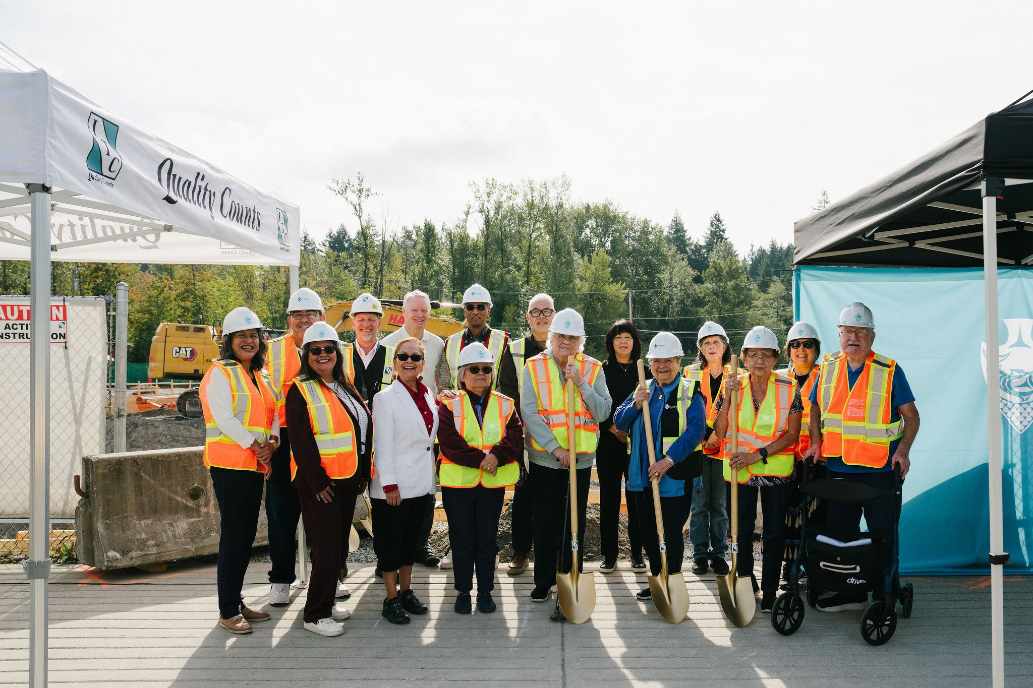 A group of attendees in hard hats and safety vests, holding ceremonial golden shovels, at the East Wind groundbreaking at leləm̓ village.