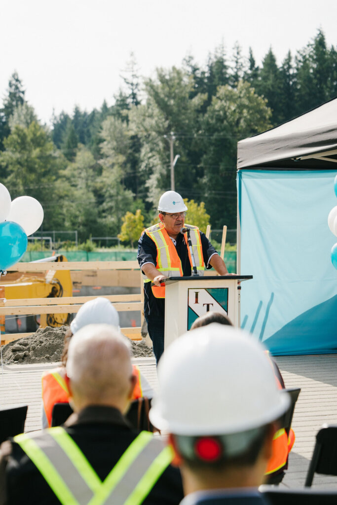 A speaker in a hard hat and safety vest at a podium addressing seated guests at the East Wind groundbreaking ceremony.