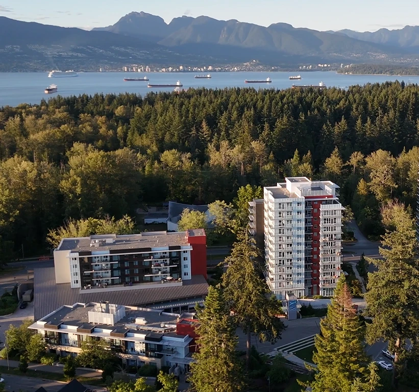 Aerial view of leləm̓ Village, with residential buildings among evergreen forest and the mountains and waterfront in the background.